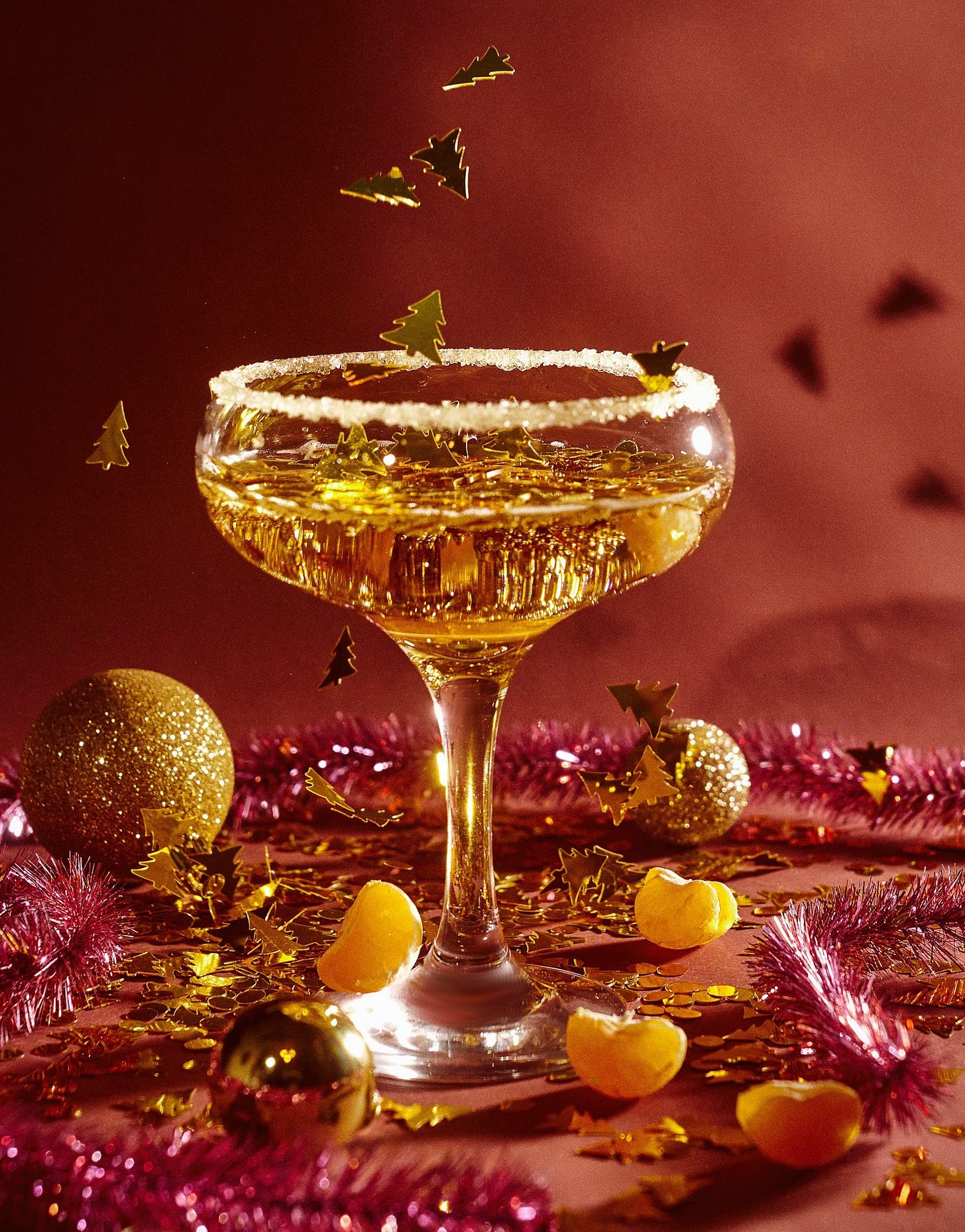 A coupe glass of champagne with sugar on the rim rests on a dark red surface with a matching red background, lit from the side. Small, flat gold Christmas tree shapes fall like confetti into and around the glass. Segments of a clementine rest among the confetti, red tinsel, and sparkly gold Christmas ball ornaments on the surface.
