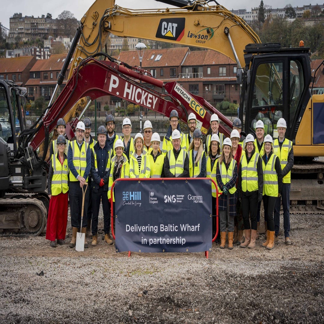 Around two dozen people in yellow hi-vis and white hard hats at a groundbreaking on cleared ground at Baltic Wharf on Bristol's harbourside, behind a banner reading "Delivering Baltic Wharf in partnership" with the Hill, Homes England, Sovereign Network Group and Goram Homes logos; two excavators and harbourside housing stand behind them. Around two dozen people in yellow hi-vis and white hard hats at a groundbreaking on cleared ground at Baltic Wharf on Bristol's harbourside, behind a banner reading "Delivering Baltic Wharf in partnership" with the Hill, Homes England, Sovereign Network Group and Goram Homes logos; two excavators and harbourside housing stand behind them.