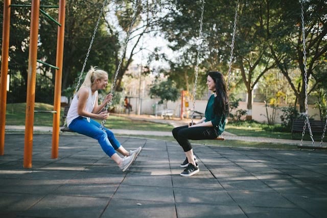 Two young women on a playground, sitting on swings they have twisted so they are facing each other.