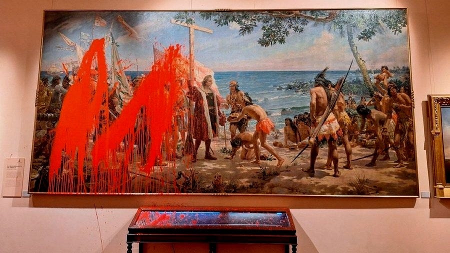 A large rectangular canvas painting portrays a historical scene of indigenous figures some standing some kneeling in a coastal setting with huts trees spears and ocean waves in the background rendered in classical style with detailed brushwork and earthy tones overlaid with chaotic orange paint drips and splatters resembling graffiti across the figures landscape and sky creating a defaced appearance the artwork is mounted on a museum wall beside a smaller framed picture and ornate gold frame with a red pedestal below and soft pink wall surroundings