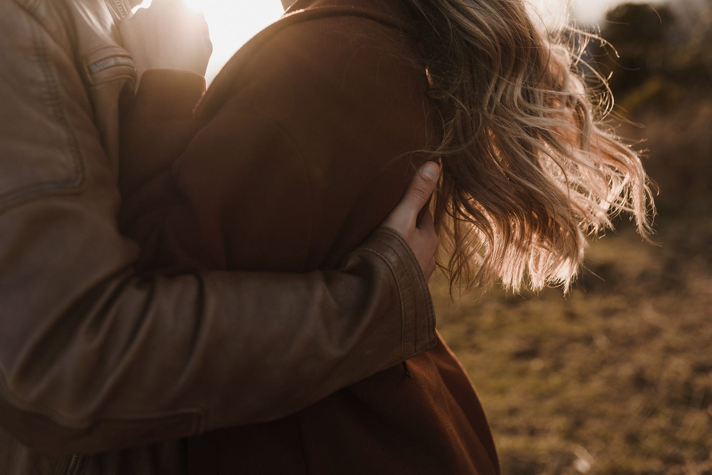 Man sieht in Nahhaufnahme eine Frau in den Armen eines Mannes, draußen bei Abendsonne. Die beiden Tragen Mäntel, die Köpfe sind vom Bild abgeschnitten. Man sieht nur die langen blonden Haare der Frau, die im Wind wehen. Man sieht in Nahhaufnahme eine Frau in den Armen eines Mannes, draußen bei Abendsonne. Die beiden Tragen Mäntel, die Köpfe sind vom Bild abgeschnitten. Man sieht nur die langen blonden Haare der Frau, die im Wind wehen.
