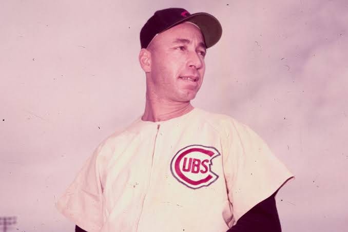 Jones is shown in color, standing but looking away from the camera with clouds on one light of a ballpark behind him. He wears a white jersey with a red Cubs logo and black baseball hat. He has a light fluorescent beige complexion, strong eyebrows, full lips and a commanding nose. 