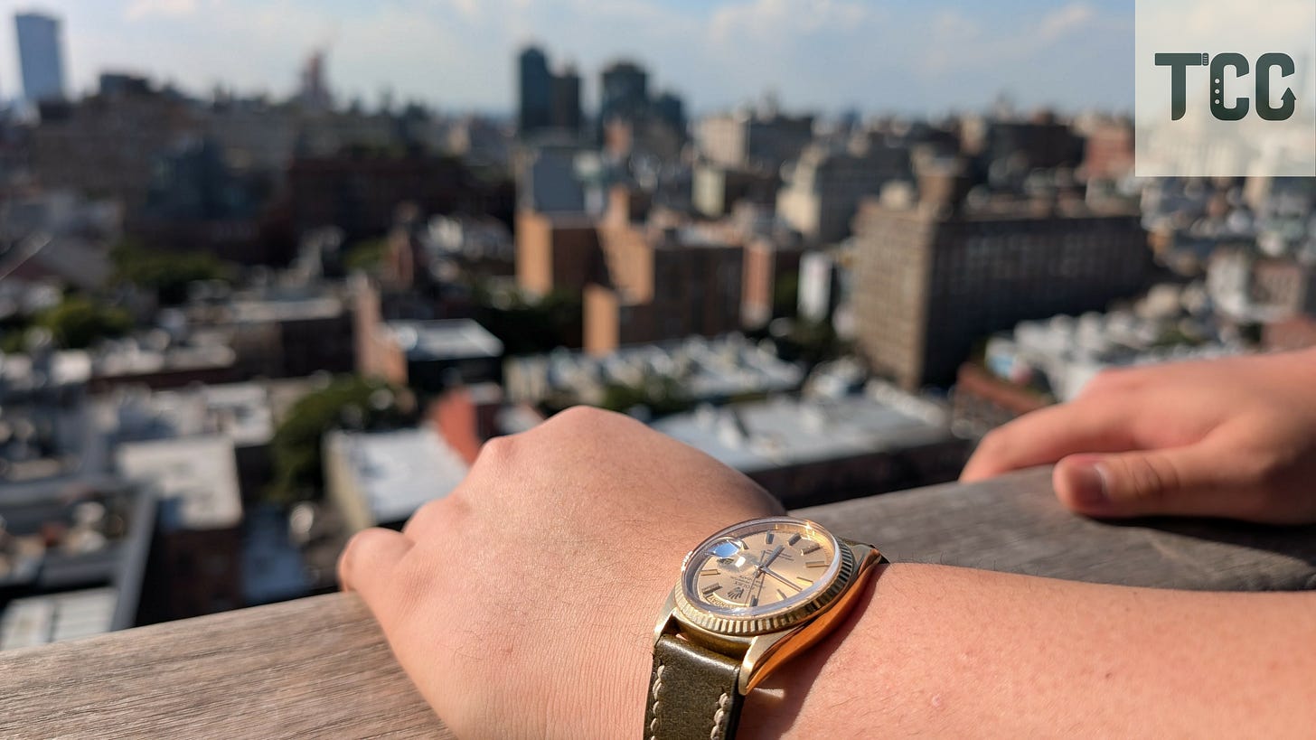 Full wrist-shot of the Rolex Day-Date with downtown New York skyline blurred behind.