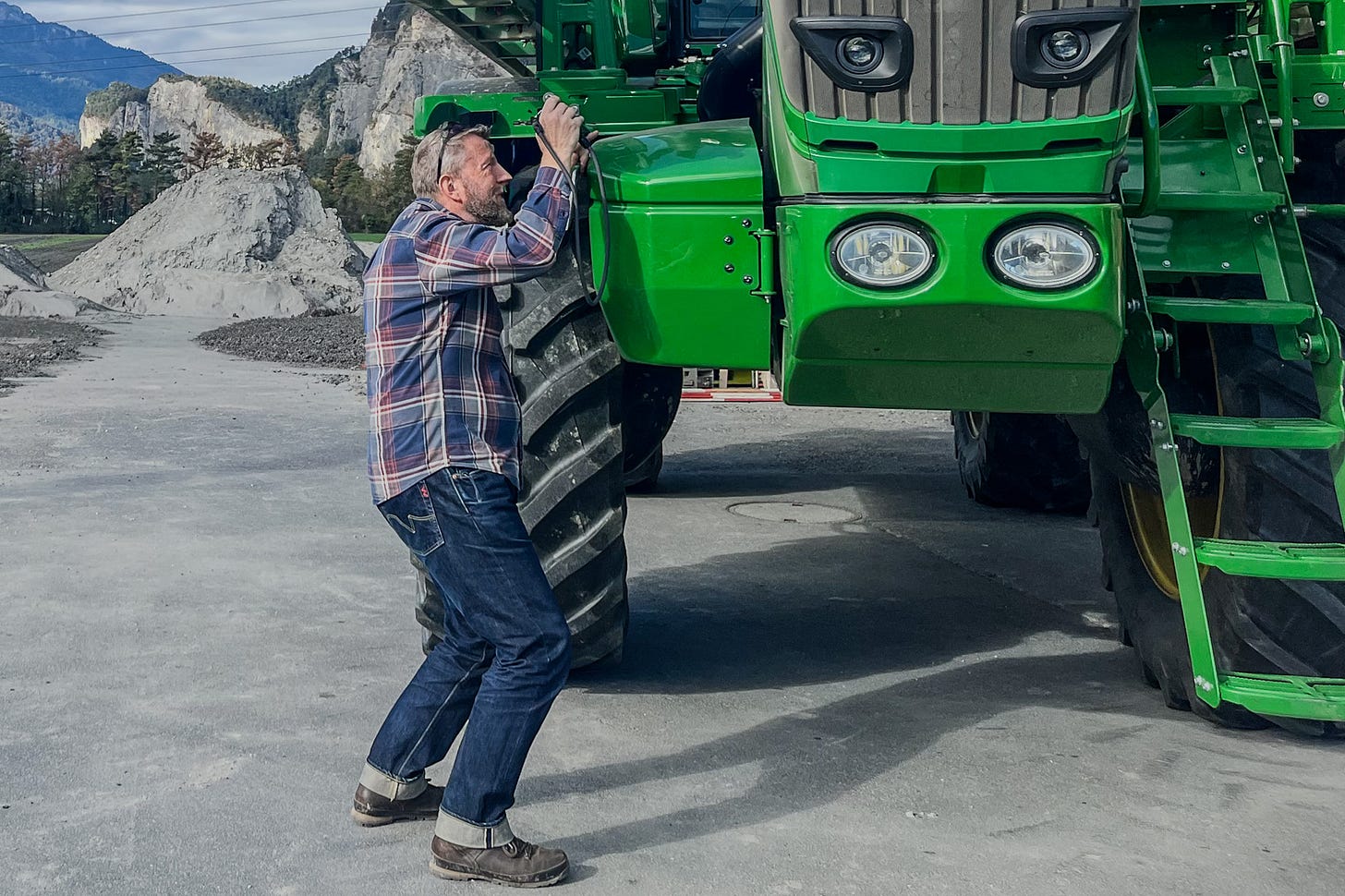Der Agrarjournalist Jürg Vollmer mit kariertem Hemd, Jeans und Arbeitsschuhen fotografiert eine riesige grüne John-Deere-Feldspritze R4140i. Die Maschine mit ihren mächtigen Reifen und den gelben Felgen überragt ihn deutlich. Im Hintergrund sind felsige Berge zu sehen. Der Agrarjournalist Jürg Vollmer mit kariertem Hemd, Jeans und Arbeitsschuhen fotografiert eine riesige grüne John-Deere-Feldspritze R4140i. Die Maschine mit ihren mächtigen Reifen und den gelben Felgen überragt ihn deutlich. Im Hintergrund sind felsige Berge zu sehen.