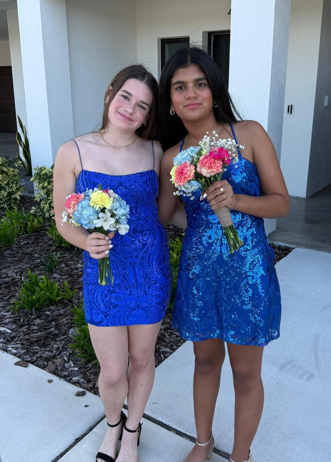 Two teenage girls, Lexie and Arianna, smiling together outdoors in blue dresses, holding a bright bouquet of pink and blue flowers with fresh, natural makeup.