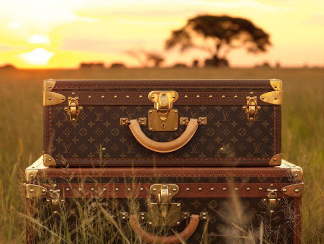 Two stacked Louis Vuitton monogram-canvas trunks with brass hardware sitting in tall savanna grass, with an acacia tree silhouetted against an orange African sunset in the background — visualizing LVMH's pitch at the 2026 Annual General Meeting positioning Africa as the next growth frontier for the group's Wines & Spirits and Fashion & Leather Goods divisions.