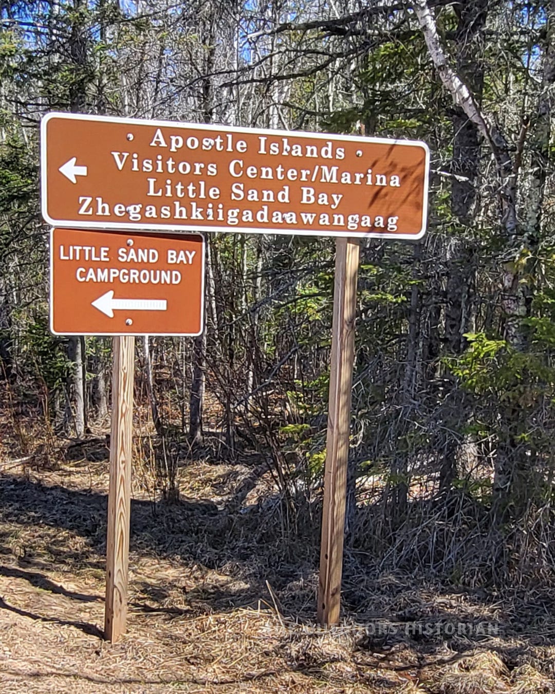 A vertical (4:5) photo of the brown and white National Park Service sign for the Apostle Islands Little Sand Bay Visitor Center. Below the English text, the Anishinaabe name "Zegashkiigadaawangaag" is clearly displayed on a sign along the wooded Little Sand Bay Road.