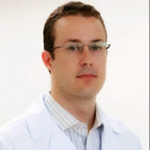 Homem de pele clara com cabelos curtos e castanhos, que usa óculos de grau finos e sem moldura. Ele está vestido com um jaleco branco aberto sobre uma camisa social branca com listras finas. Sua expressão facial é neutra. O fundo é simples, branco e liso, sem quaisquer distrações visuais. Homem de pele clara com cabelos curtos e castanhos, que usa óculos de grau finos e sem moldura. Ele está vestido com um jaleco branco aberto sobre uma camisa social branca com listras finas. Sua expressão facial é neutra. O fundo é simples, branco e liso, sem quaisquer distrações visuais.