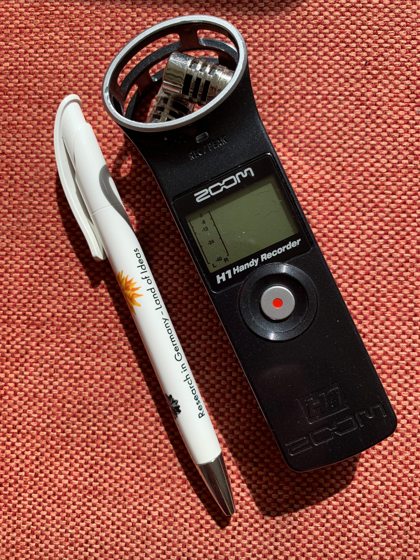 A hand-held digital recorder laid against orange upholstery. Next to it is a pen for scale (it is a similar length to the pen, and about 5 centimetres wide).