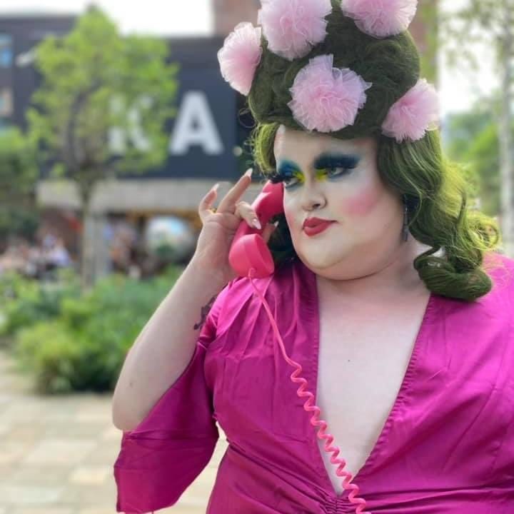 A white drag queen wears a pink robe with tall hair studded with plumes of pink fabric, holding a pink wired landline phone to her ear.