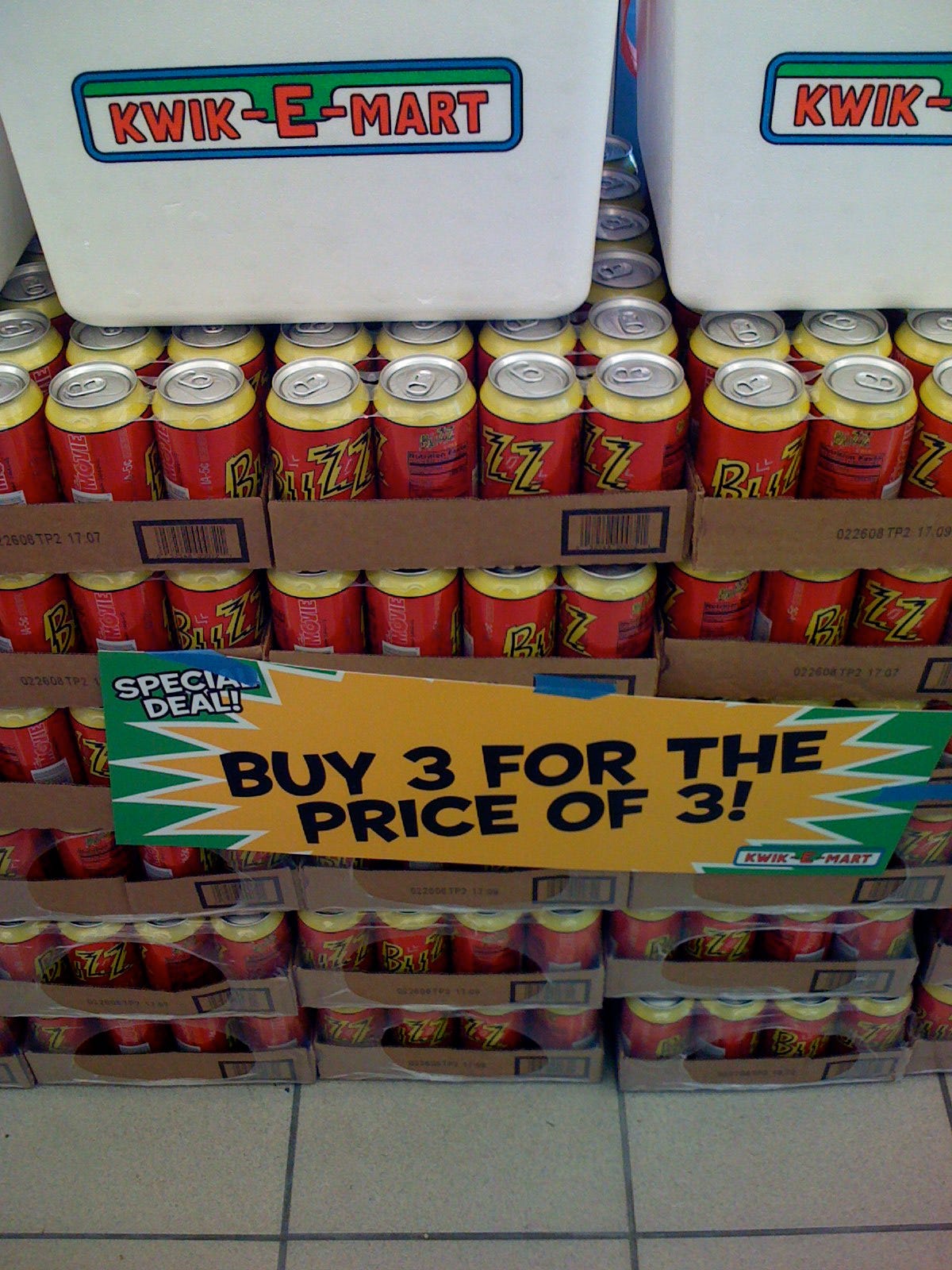 Store display of Buzz Cola with Kwik-E-Mart logoed coolers. A printed sign on the pallets of cola reads "Special Deal! Buy 3 for the price of 3!" The Kwik-E-Mart logo also appears on the sign.