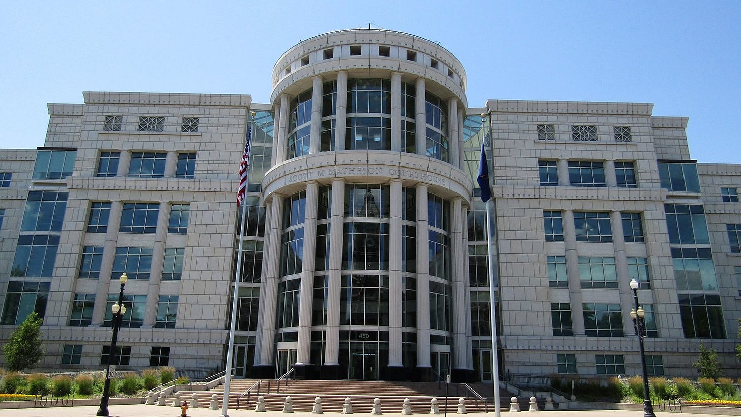 A tall cream-colored stone courthouse with a columned glass rotunda at its center, wide entrance steps and a U.S. flag out front under a clear blue sky. A tall cream-colored stone courthouse with a columned glass rotunda at its center, wide entrance steps and a U.S. flag out front under a clear blue sky.