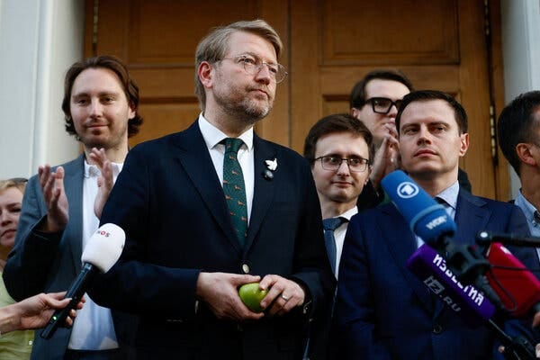 A group of people, many in suits and ties, stand before colorful microphones outside a wooden door. A bespectacled person at center holds a green apple.