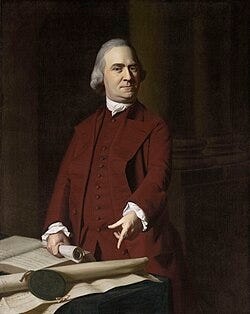 A stern middle-aged man with gray hair is wearing a dark red suit. He is standing behind a table, holding a rolled up document in one hand, and pointing with the other hand to a large document on the table.
