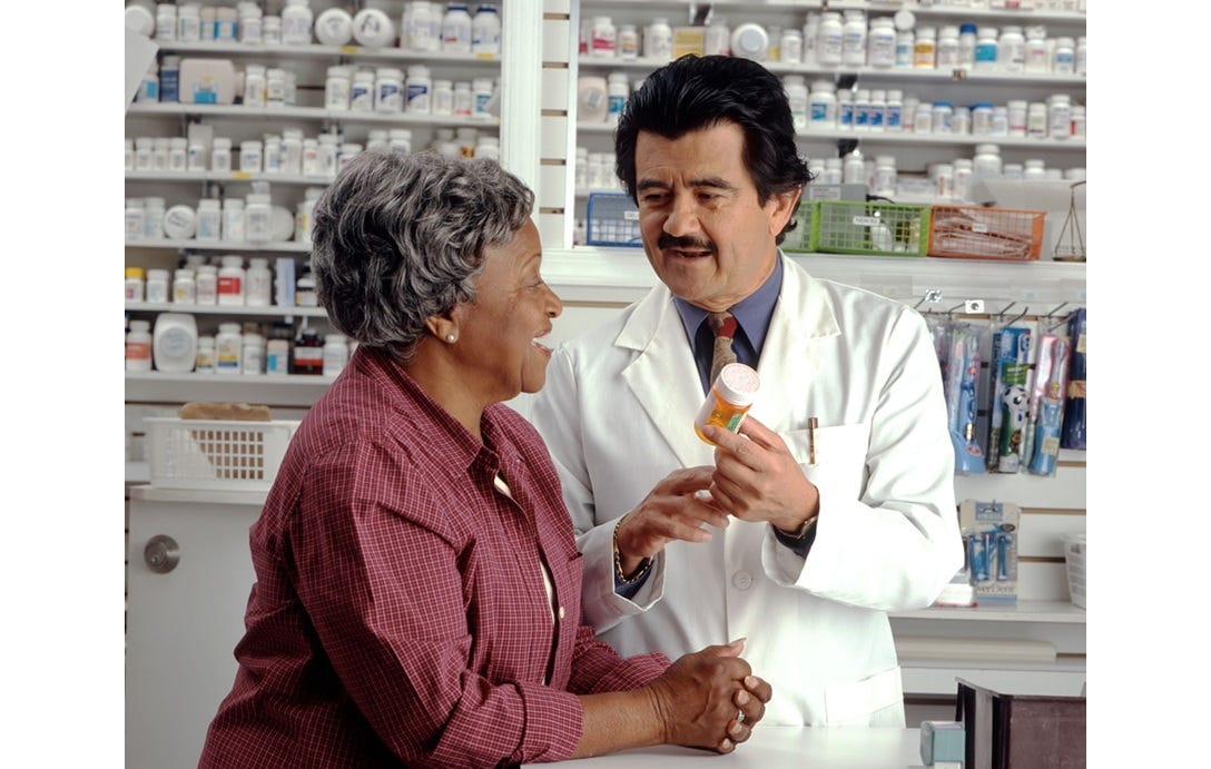 In a pharmacy, an elderly Black woman talks to a Hispanic pharmacist wearing a white lab coat. He is showing her a bottle of pills. In a pharmacy, an elderly Black woman talks to a Hispanic pharmacist wearing a white lab coat. He is showing her a bottle of pills.