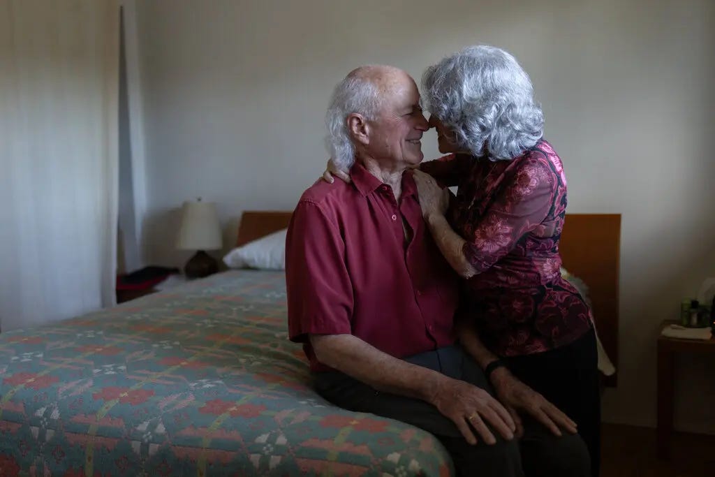 Joan Price, standing, touches noses with Mac Marshall, who is sitting on the side of a bed and wearing a red shirt. They are both smiling.