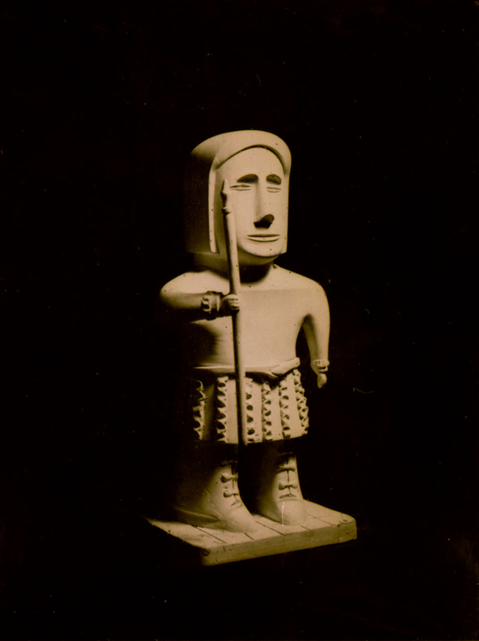 A imagem exibe uma escultura, que é uma obra do artista brasileiro autodidata Lúcio Noeman.  Assunto: A escultura retrata uma figura humana estilizada, descrita como um dos "guerreiros empenhados na luta entre o bem e o mal" que o artista frequentemente criava.  Características Físicas: A figura tem uma aparência robusta, com pernas curtas e grossas, e usa botas. Ela veste uma espécie de saia ou cinto com textura detalhada e segura um objeto longo, semelhante a um cajado ou lança, com a mão esquerda.  Estilo e Expressão: O rosto é alongado, com traços simplificados e uma expressão séria ou melancólica. A cabeça é coberta por um capuz ou capacete arredondado. O estilo da obra é notável pela sua qualidade artística e profundidade emocional.  Iluminação: A fotografia utiliza uma iluminação dramática, com um fundo escuro que destaca a textura e a forma da escultura, que parece ser feita de barro ou material similar. 