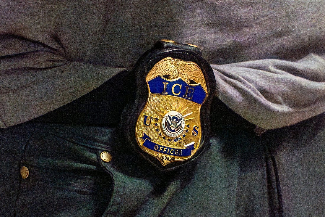An ICE badge is on the waist of someone wearing a very sloppily tucked-in gray athletic tee and baggy black pants.