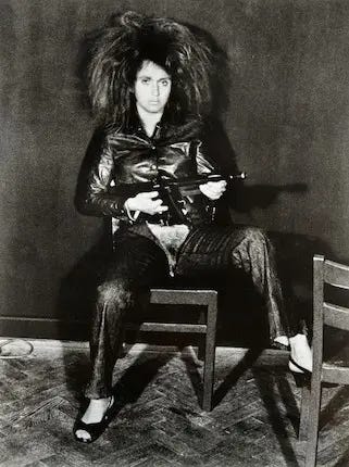 Black-and-white photograph of VALIE EXPORT seated on a chair wearing black leather pants with the crotch cut away, exposing her pelvis. She holds a machine gun across her lap and stares directly at the camera while two empty chairs frame the foreground. Black-and-white photograph of VALIE EXPORT seated on a chair wearing black leather pants with the crotch cut away, exposing her pelvis. She holds a machine gun across her lap and stares directly at the camera while two empty chairs frame the foreground.