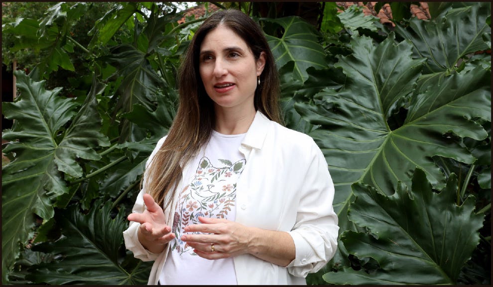Mulher adulta em ambiente externo, diante de grandes folhas verdes que lembram um jardim tropical. Ela tem cabelos longos castanhos, usa brincos pequenos e veste uma camisa branca aberta sobre uma camiseta clara com estampa floral. Está com as mãos levemente erguidas, como se estivesse explicando algo, e apresenta expressão atenta e envolvida. A iluminação é natural e suave.