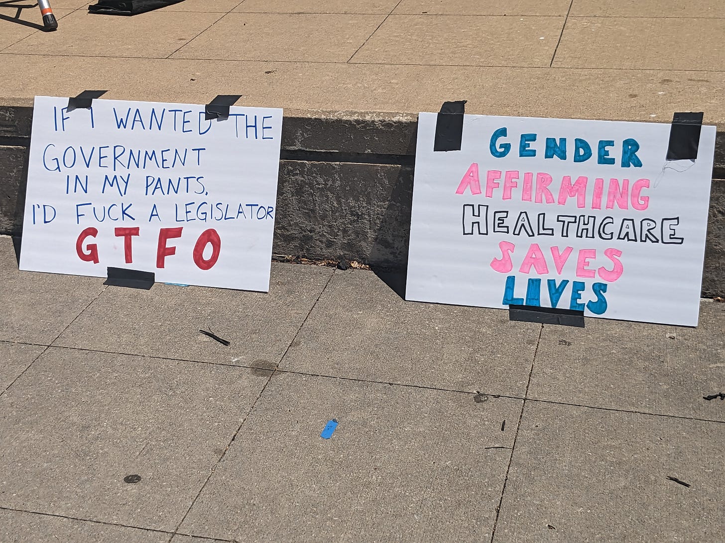 A photograph of two signs taped to the edge of a stage. One reads: "If I wanted the government in my pants, I'd fuck a legislator. GTFO" The other says: "Gender Affirming Healthcare Saves Lives"