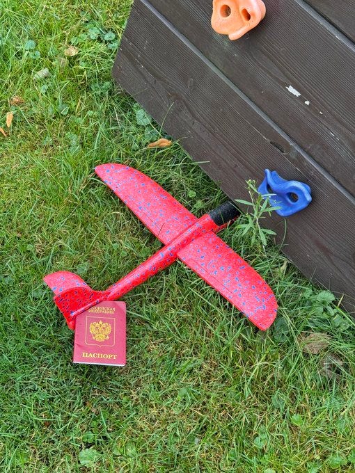 A red toy airplane with a pattern, lying on grass near a wooden structure with blue and orange climbing holds. A red Russian passport is placed beside the airplane. Text on the airplane and passport is visible.