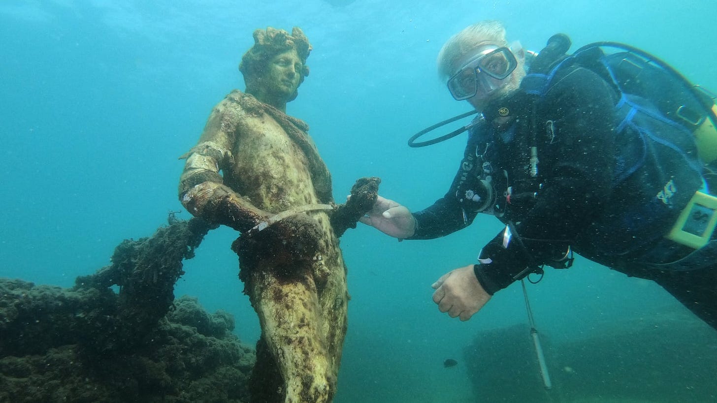 A dive guide shows tourists a copy of the original statues preserved at the Museum of Baiae, representing Dionysus with ivy crown in the Nymphaeum of punta Epitaffio, the submerged ancient Roman city of Baiae at the Baiae Underwater Park, part of the Campi Flegrei Archaeological Park complex site in Pozzuoli near Naples.