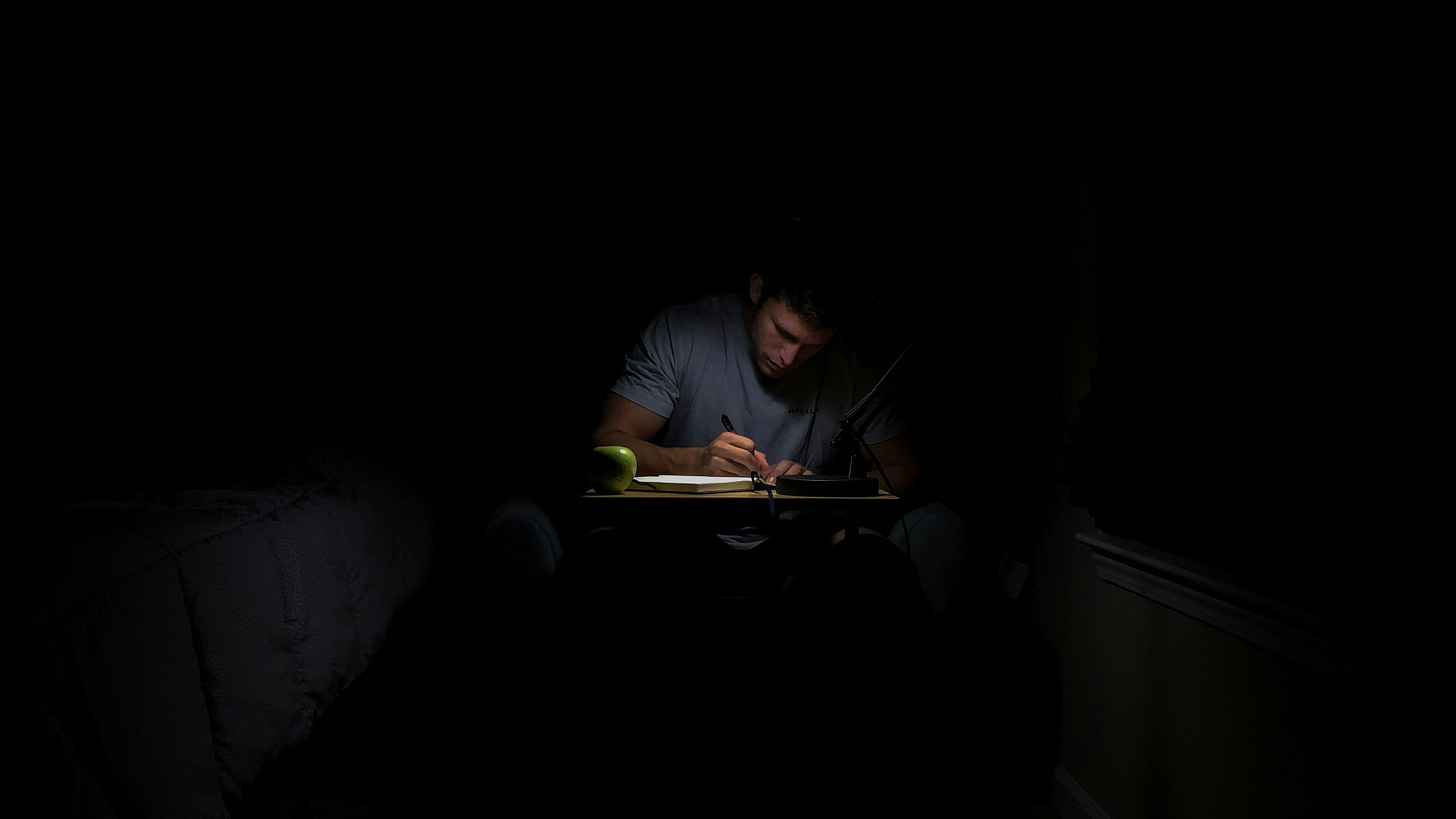 A barely visible man writes at a desk, surrounded by darkness A barely visible man writes at a desk, surrounded by darkness