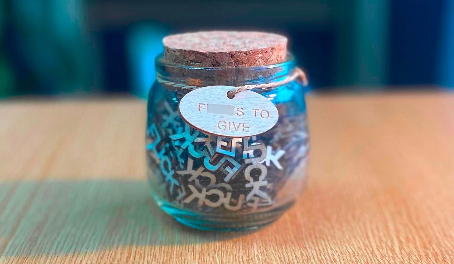 A glass jar with a cork lid sitting on a desk. It is labelled "F*cks to Give" and inside are lots of laser cut pieces which each spell F*CK