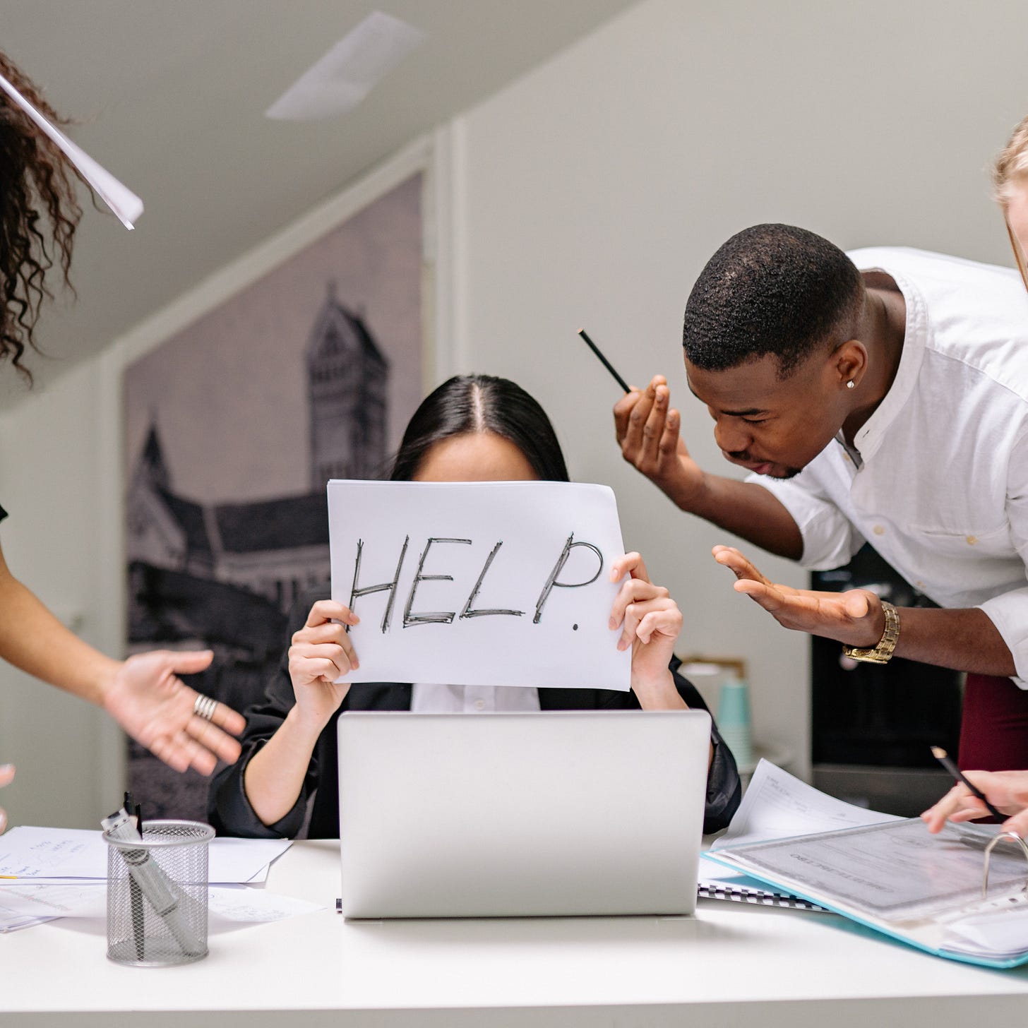 a person with long hair is hiding behind a paper, holding up the word HELP written on it as other team members yell at her