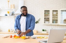 A focused looking person chops colorful vegetables on a kitchen counter in a modern kitchen while looking toward an open laptop that sits on the counter.