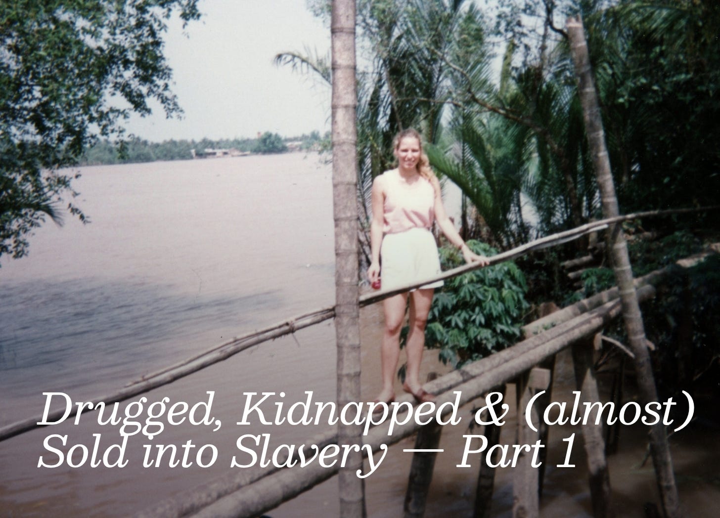 A young white woman with long blond hair is wearing a pink top and short while standing on a bamboo bridge. The is brown water in the river underneath and jungle along the sides.