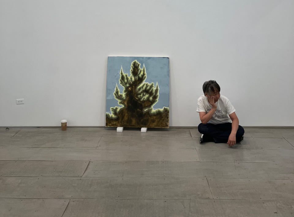 An Asian man seated cross-legged on the floor beside a painting of a tree resting on two boards. A coffee cup is next to the painting on the floor.