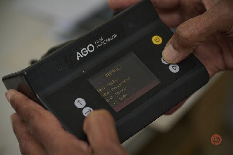 Close-up of hands holding an AGO film processor with menu options displayed on its screen.