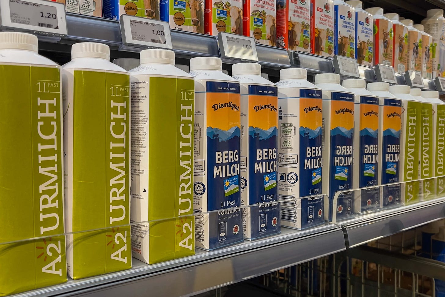 Das Foto zeigt die «A2 Urmilch» und die «Diemtigtaler Bergmilch» aus der Molkerei und Käserei «Simmental Switzerland» im Kühlregal einer Migros-Filiale. Die Milchprodukte sind in Tetrapaks abgefüllt und in zwei Reihen nebeneinander angeordnet. Die grünen Verpackungen der «A2 Urmilch» heben sich von den blauen Verpackungen der «Diemtigtaler Bergmilch» mit einer Berglandschaft und einer Schweizer Flagge ab. Darüber sind weitere Milchprodukte verschiedener Marken sichtbar, ebenfalls in einem gut beleuchteten Kühlregal präsentiert. Die Preisschilder zeigen die Produkte als Teil des regionalen Sortiments.