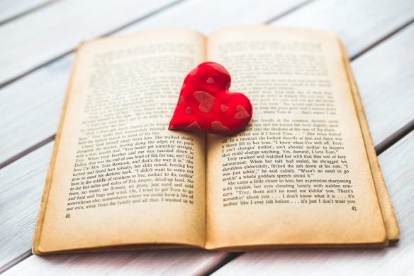 This image shows an open book laying flat on a white surface. There is a red heart on top of the book in the crease between the two pages. This image shows an open book laying flat on a white surface. There is a red heart on top of the book in the crease between the two pages.