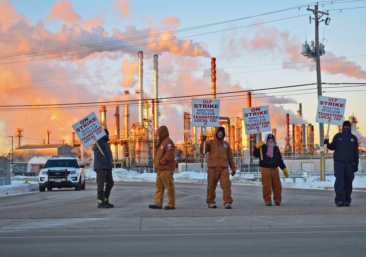 picketers stand in the road in front of an oil refinery sending clouds of smoke reflecting orange and purple in the the blue sky. their signs read "ULP strike marathon petroleum teamsters local 120" picketers stand in the road in front of an oil refinery sending clouds of smoke reflecting orange and purple in the the blue sky. their signs read "ULP strike marathon petroleum teamsters local 120"