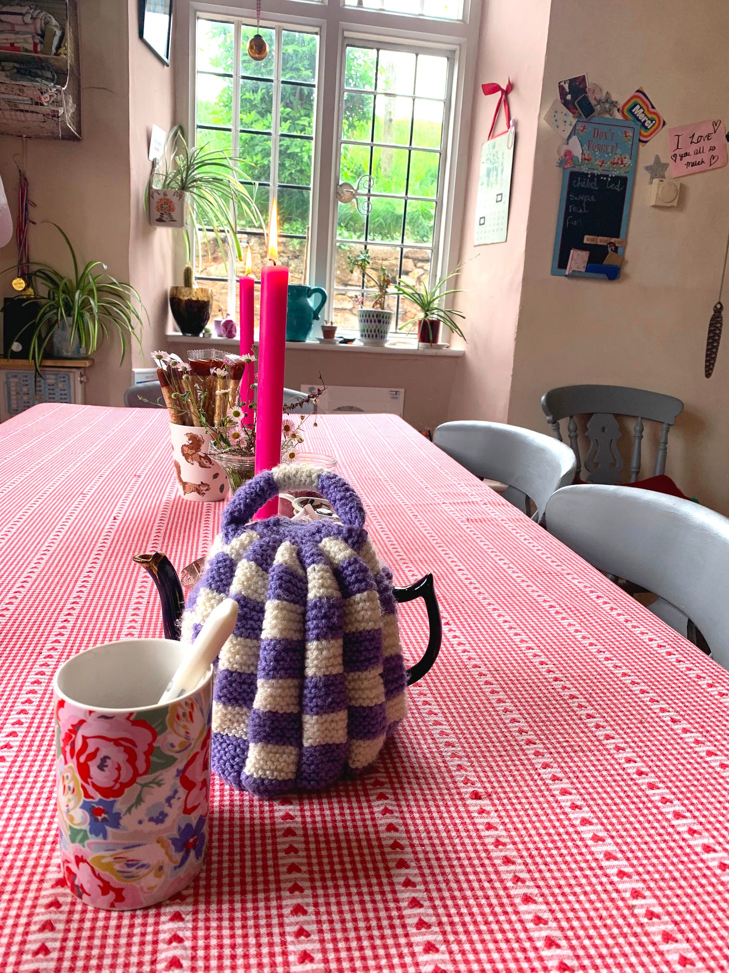 A crocheted teapot & floral mug on a long table in a lived-in kitchen, with candles and a window overlooking green fields