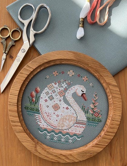 A finished cross stitch project in a round, wooden frame.  The cross stitch is of a swan, floating in water, with detail work within the feathers, water waves, and landscape.