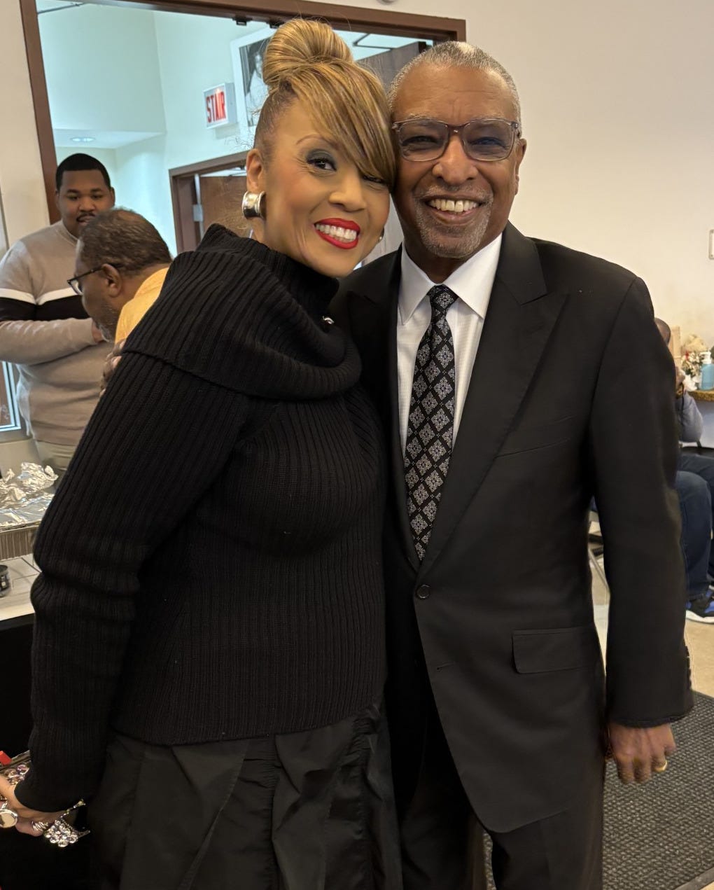 Santita Jackson, wearing a black turtleneck sweater, black skirt, and round silver earrings smiles while posing in a side hug with Dr. Soaries, who is wearing a black suit, white shirt, black and silver patterned tie, and glasses. He is also smiling.