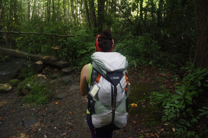 Two images of Nicole in Smoky Mountain National Park. In the first, she poses in front of the park sign in Cataloochee (Eastern Tennessee). In the second image, we see the back of Nicole's hiking pack as she faces away toward a wooden bridge crossing a creek.