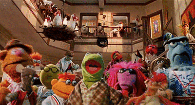 "Un'ampia inquadratura di numerosi Muppets che guardano avanti mentre si trovano sulle scale e sul pianerottolo di un'abitazione, con Kermit la Rana in primo piano al centro. Sopra c'è un lampadario decorato. "Un'ampia inquadratura di numerosi Muppets che guardano avanti mentre si trovano sulle scale e sul pianerottolo di un'abitazione, con Kermit la Rana in primo piano al centro. Sopra c'è un lampadario decorato.