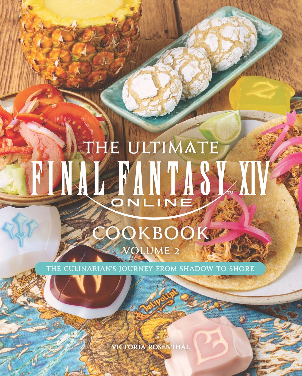 Cover of The Ultimate Final Fantasy XIV Online Cookbook: Volume 2 – The Culinarian’s Journey from Shadow to Shore by Victoria Rosenthal, featuring tropical-themed dishes like tacos, a tomato salad, pineapple drink, and powdered cookies arranged over a map of Eorzea. Colorful class crystal replicas are scattered across the scene, tying into the game’s world.