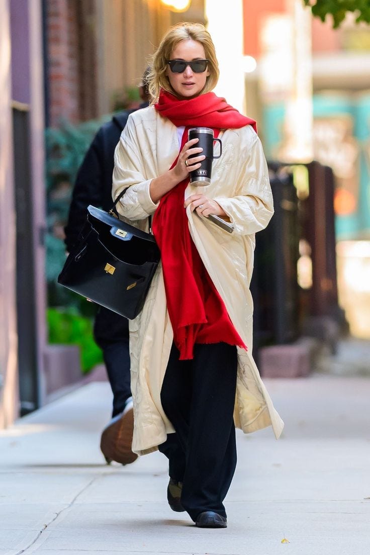 This may contain: a woman walking down the street while holding a coffee cup and wearing a red scarf