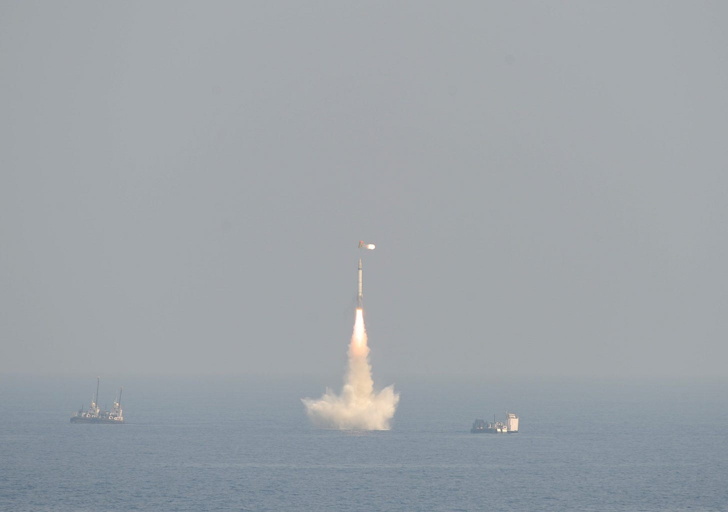 A missile rises from the sea on a column of flame and spray, with two support vessels on the horizon
