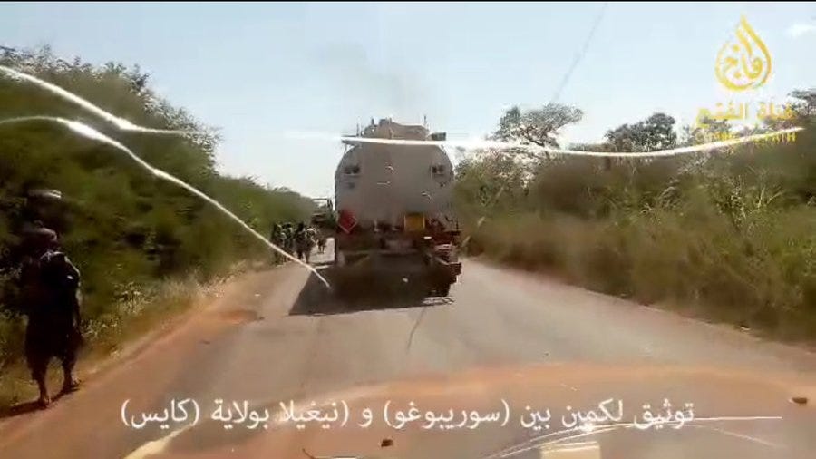 A white tanker truck travels on a dirt road flanked by green bushes and trees under a clear sky with two individuals in dark clothing standing nearby and a yellow logo visible on the truck.