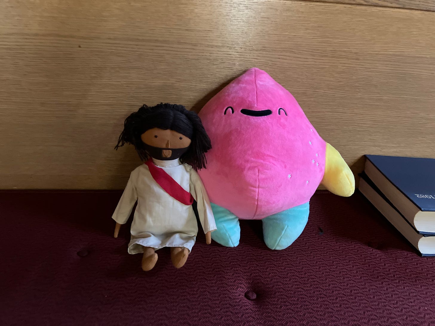A small stuffed Jesus doll in a white robe and red sash and a pink flame-shaped plush with limbs and a smiling face sit next to each other on a pew beside a small stack of hymnals.