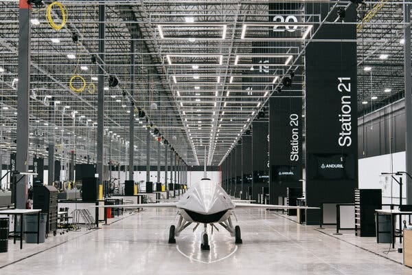 A white, sleek, space-age-looking aircraft sits on a long factory hallway with a shiny white floor.