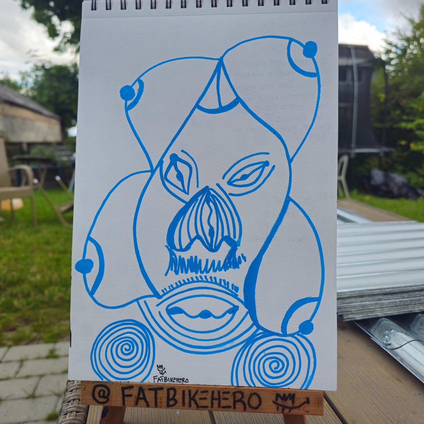 Front: A vertical blue marker drawing on a sketchbook page by artist Fatbikehero. The image features an abstract, surreal face titled "Strange Man" with spiral circles at the base and bulbous, organic facial features.