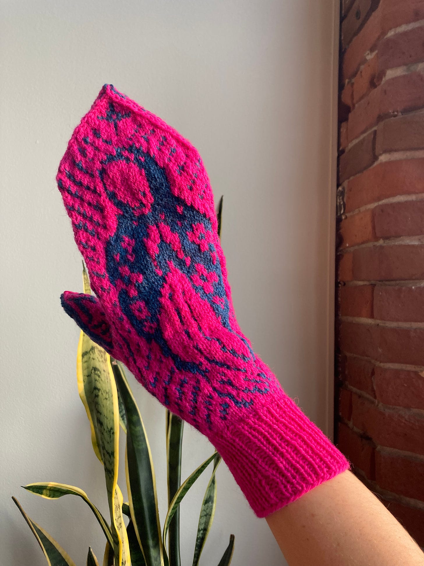 A hot pink and teal blue handknit mitten is on a person's hand. The colorwork mitten shows Mother Mary depicted as Our Lady of Guadalupe.